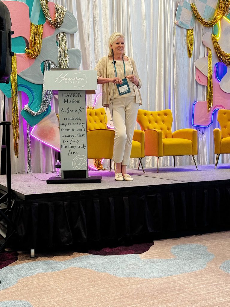 A woman stands on a stage next to a podium with a sign that reads, "Haven's Mission: connect & empower creatives," and colorful decorations in the background.