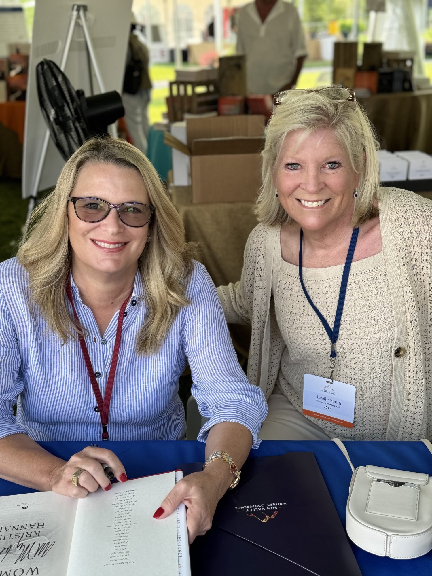 Two women smiling at a table; one is seated holding an open book, while the other stands beside her. Both are wearing name badges and appear to be attending an event.