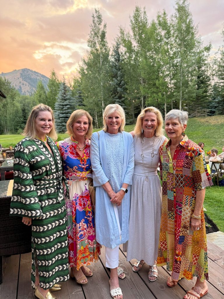 Five women stand together outdoors on a wooden deck, smiling at the camera. They are dressed in colorful, patterned clothing. There are trees and a mountain in the background.