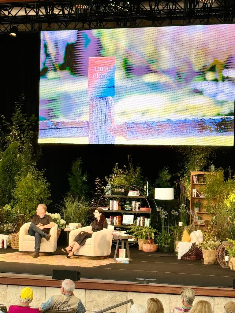 A man and a woman are seated on a stage for a talk, surrounded by plants, bookshelves, and decor. An audience watches, with a large screen behind them displaying a book cover titled "atomic effect".