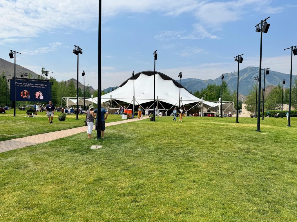 A large white tent structure set up on a green lawn with people nearby. Mountains and a partly cloudy sky are visible in the background.