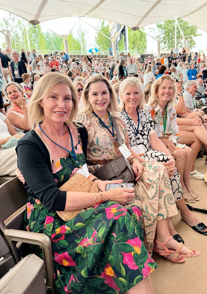 Four women seated in a covered outdoor venue, smiling at the camera with a crowd visible in the background.