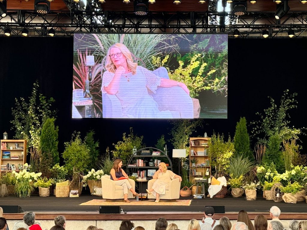 Two women are seated onstage in armchairs, having a discussion. Behind them, a large screen shows a close-up of one of the women. The stage is surrounded by bookshelves, plants, and greenery.