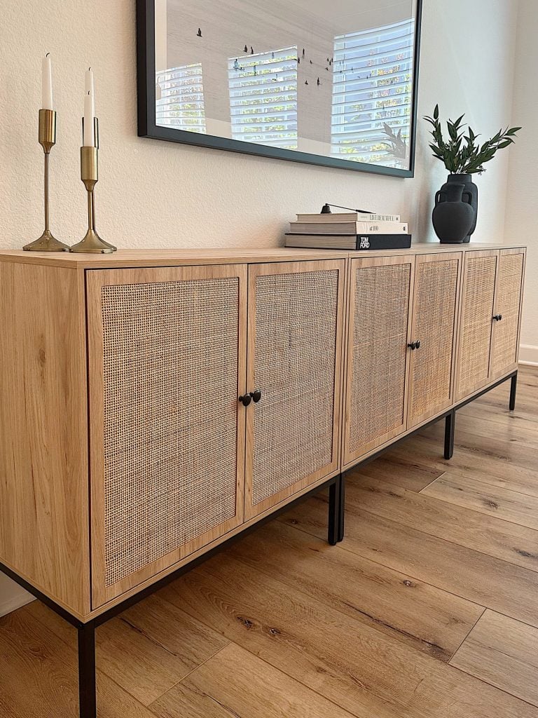 A wooden sideboard with four woven front doors, black handles, and metal legs is adorned with two candle holders, a stack of books, and a dark vase with green foliage. A framed picture hangs above.