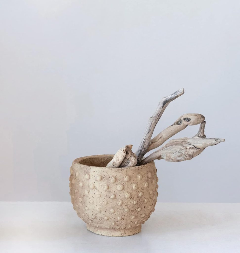 A textured beige ceramic planter holds several pieces of pale, twisted driftwood against a plain light background.