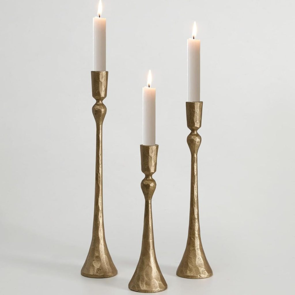 Three lit white candles in tall, gold-colored metal candle holders of varying heights create a warm, inviting look—perfect for fall room decor on a plain background.