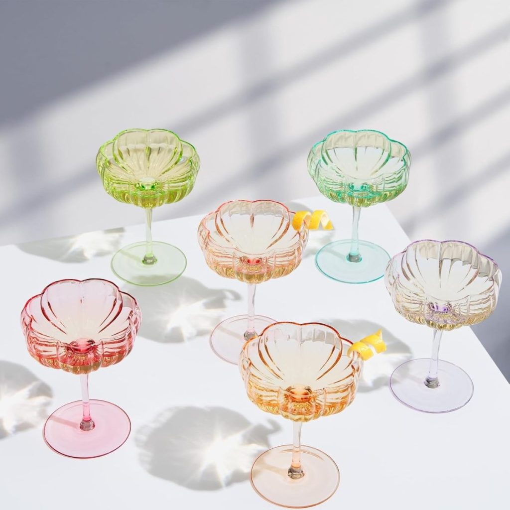 Six colorful glass goblets with flower-shaped bowls, including a striking cranberry glass, are arranged on a white surface, casting distinct shadows; two glasses contain drinks garnished with lemon twists.
