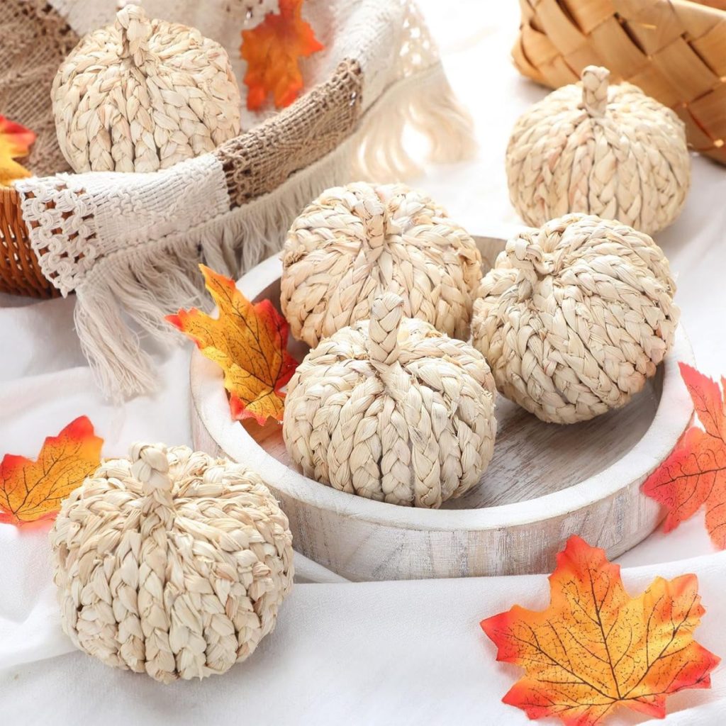 Five woven straw pumpkins in a white bowl, surrounded by artificial autumn leaves and a woven basket on a white cloth background—perfect for cozy fall home decor.