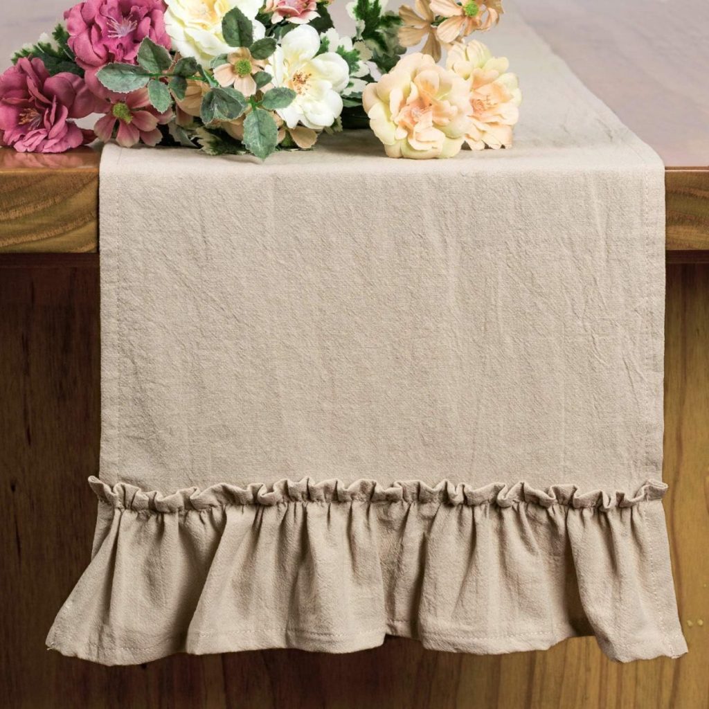 A beige fall table runner with a ruffled edge drapes over a wooden table, topped with an arrangement of artificial flowers.