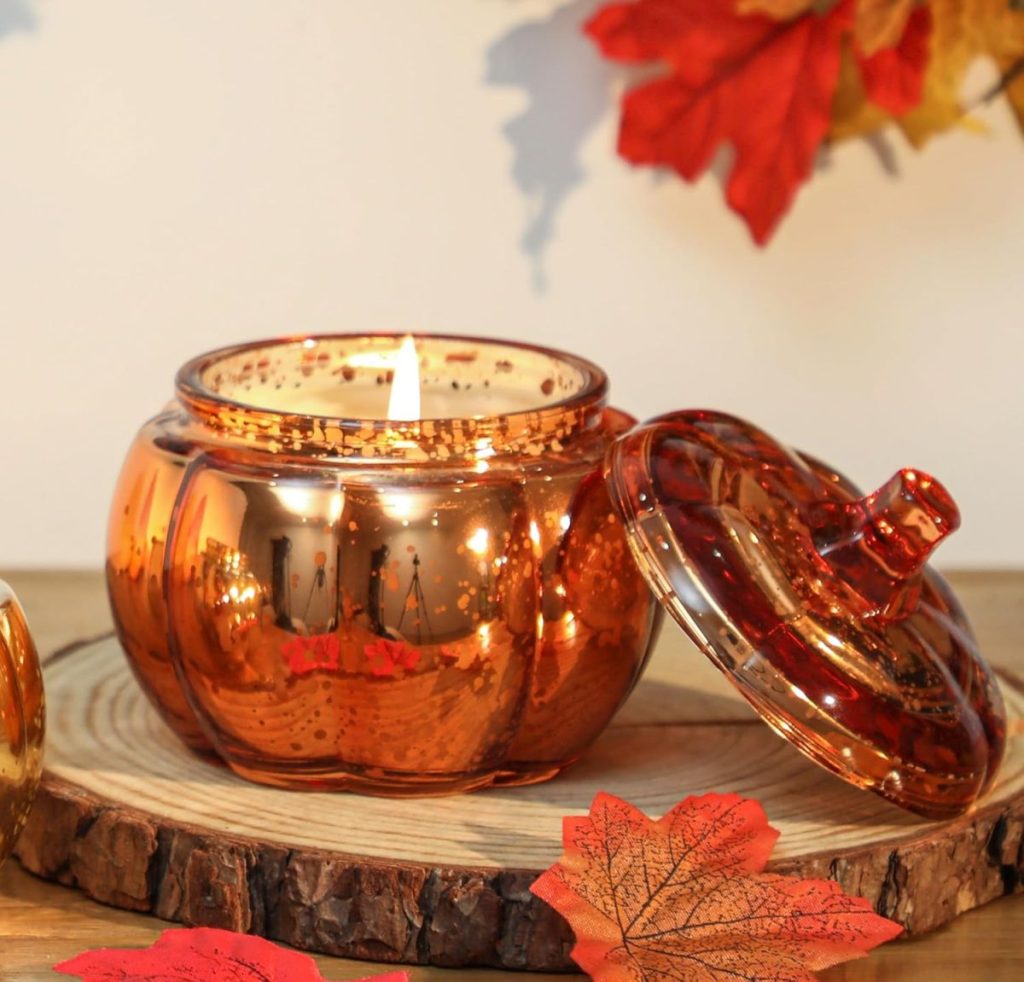 A lit candle in a pumpkin-shaped glass container with the lid off sits on a wooden slab, surrounded by autumn leaves—perfect for cozy fall home decor.