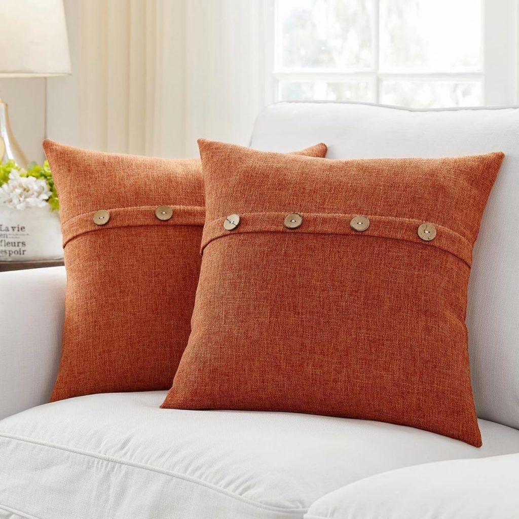 Two orange throw pillows with button accents, perfect for fall room decor, are placed on a white upholstered sofa in a bright living room.