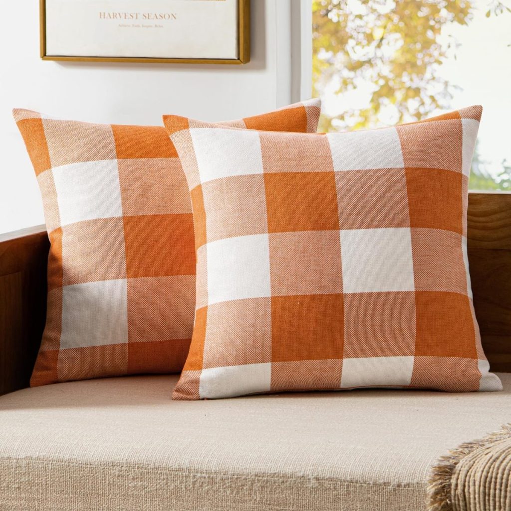 Two orange and white plaid throw pillows, perfect for fall room decor, are placed on a beige cushioned bench with a wooden backrest, near a window with sunlight streaming in.