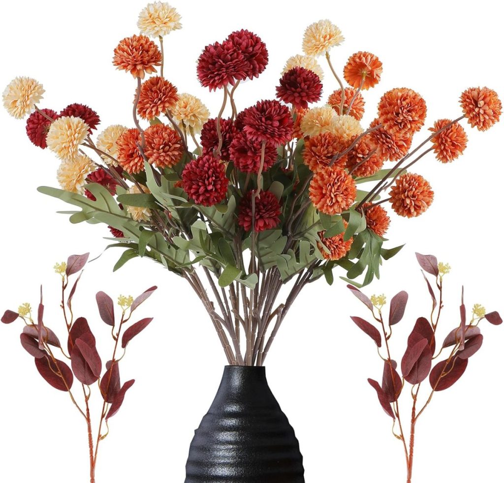 A black vase holds artificial red, orange, and cream pompom flowers with green leaves, flanked by two smaller stems with maroon leaves and yellow buds—perfect for fall home decor.