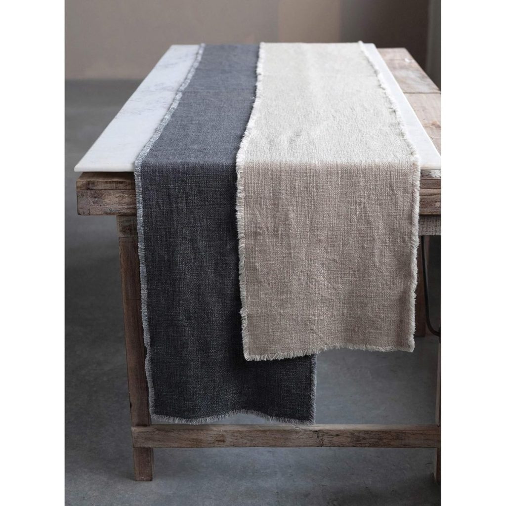 A rustic wooden table with a marble top features two linen fall table runners, one dark gray and one off-white, both with frayed edges.