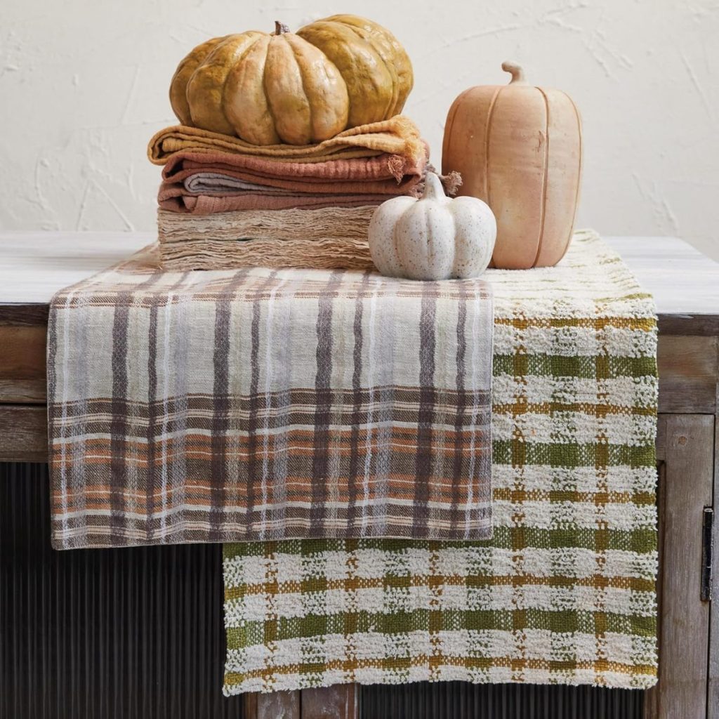 Two fall table runners draped over a rustic table with stacked folded cloths and three decorative pumpkins arranged on top.