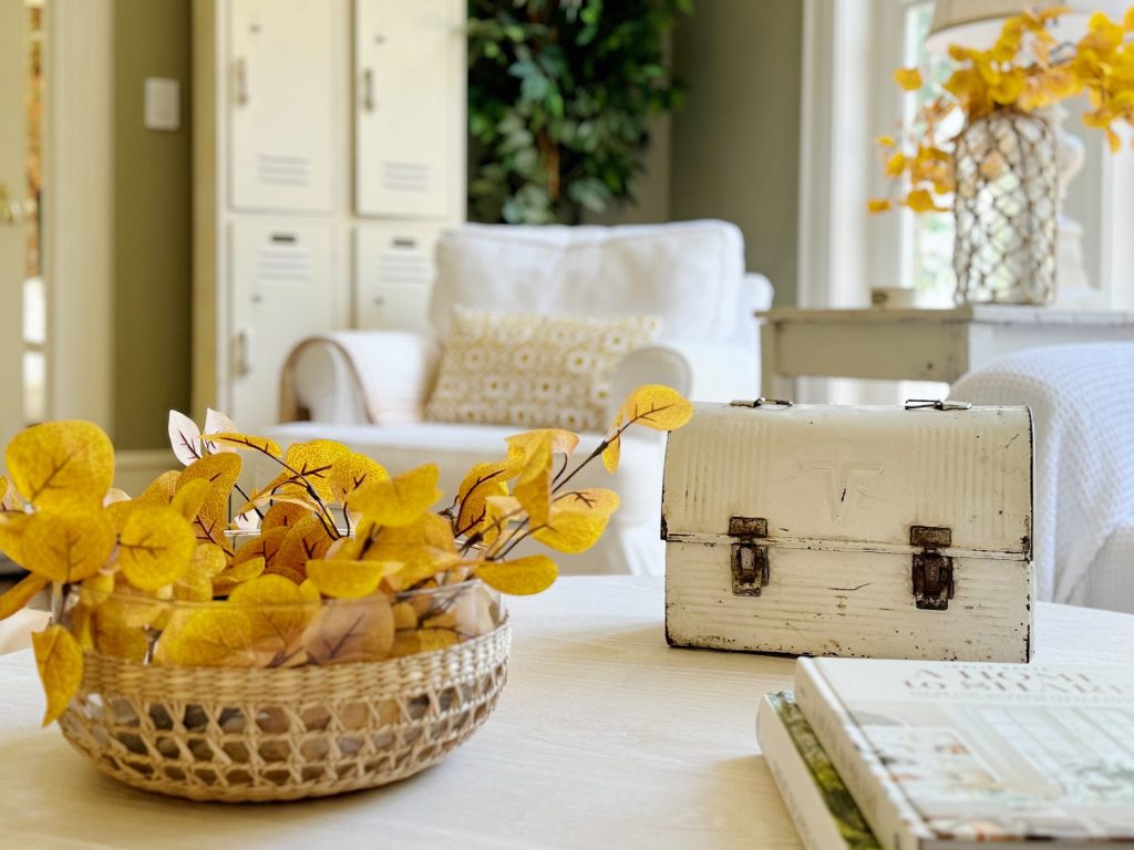 A white metal lunchbox sits on a table next to a wicker basket with yellow leaves, a stack of books, and a white armchair in the background.