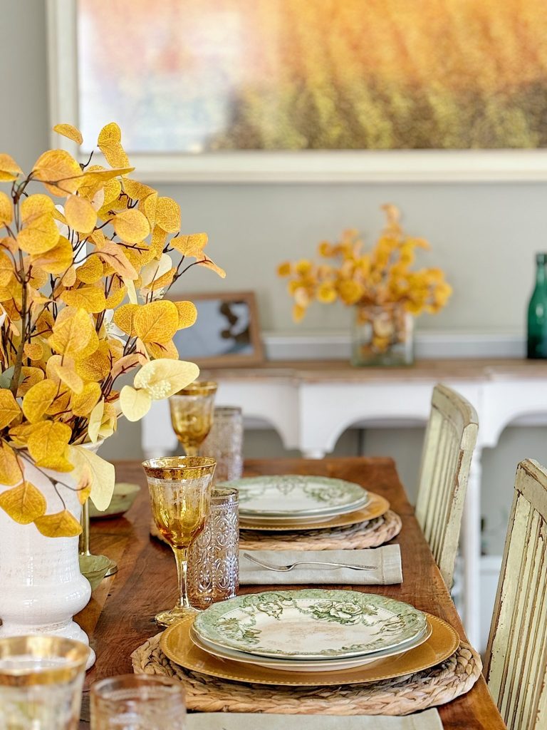 A wooden dining table set with green and white plates, wicker placemats, and yellow floral arrangements. Yellow glassware complements the rustic decor. A side table with another bouquet is in the background.