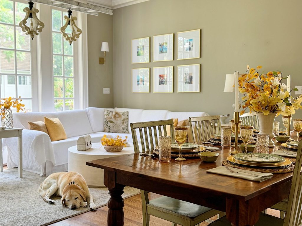 A dining and living area featuring a wooden dining table set for a meal, a white couch with pillows, framed pictures on the wall, and a dog lying on the floor near the table.