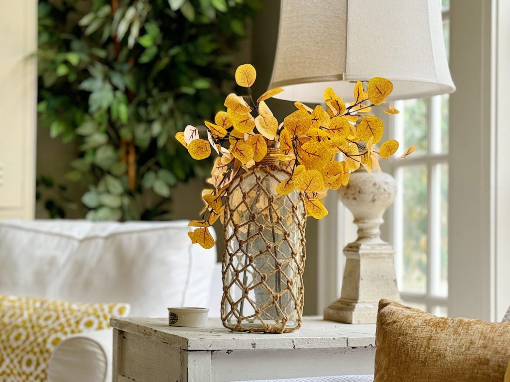 A glass vase with orange-yellow leaves sits on a rustic white side table next to a white lamp in a cozy living room with a white sofa and plush cushions. A plant is visible in the background.