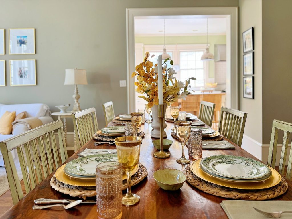 A dining table is set for a meal with plates, glasses, cutlery, and a centerpiece of yellow flowers in a vase. The background shows a living area and a kitchen.