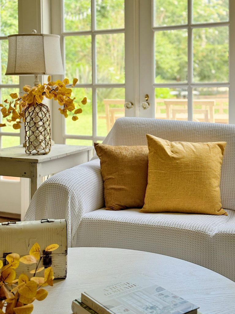 A bright living room features a white sofa with two yellow pillows, a lamp with a woven base, and a small side table. Large windows in the background overlook a green outdoor space.