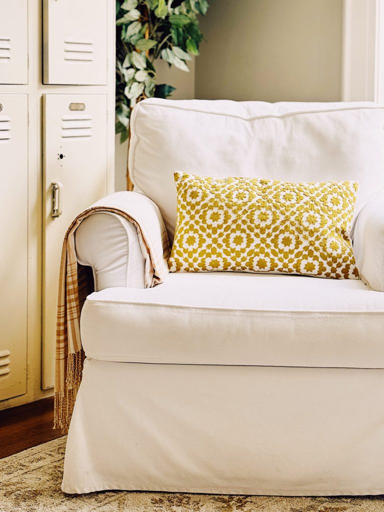 A white armchair with a yellow patterned cushion and plaid blanket sits next to vintage lockers and a potted plant.