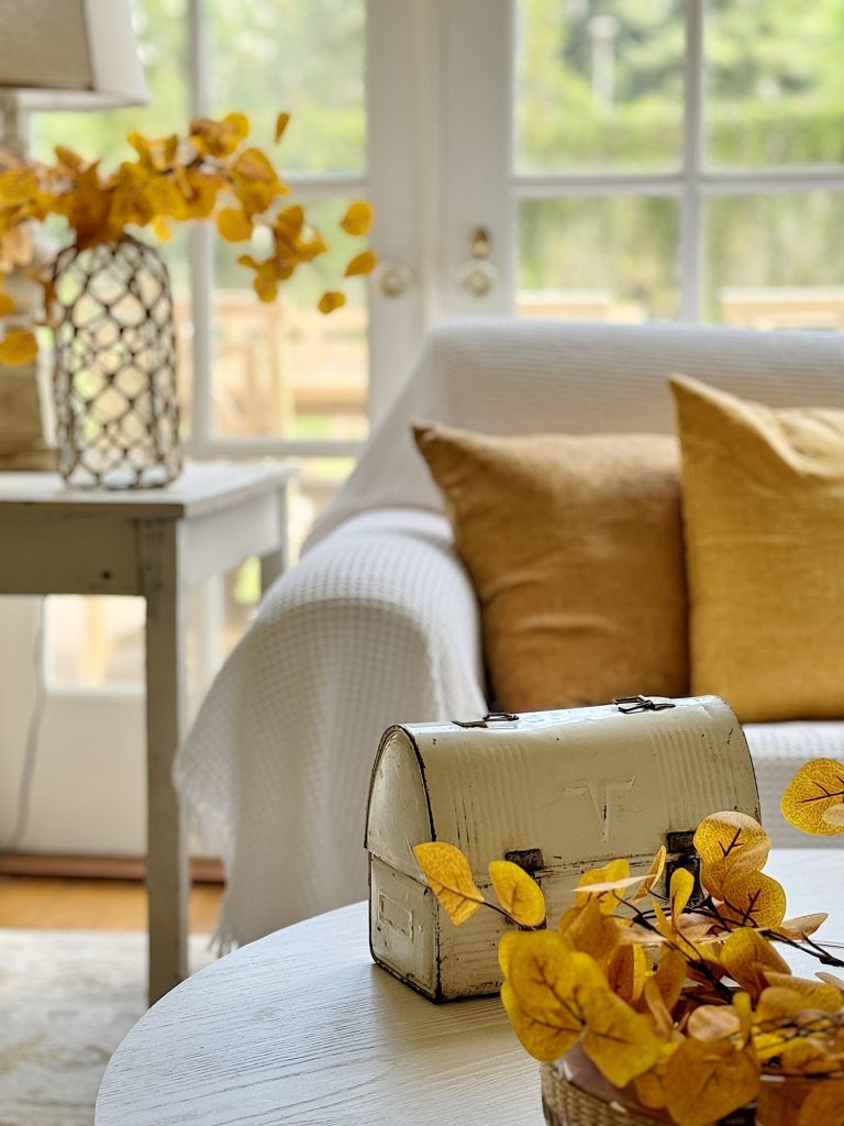 A white living room with a couch, yellow pillows, table, lantern, and autumnal leaves decorating the space; light floods in through French doors.