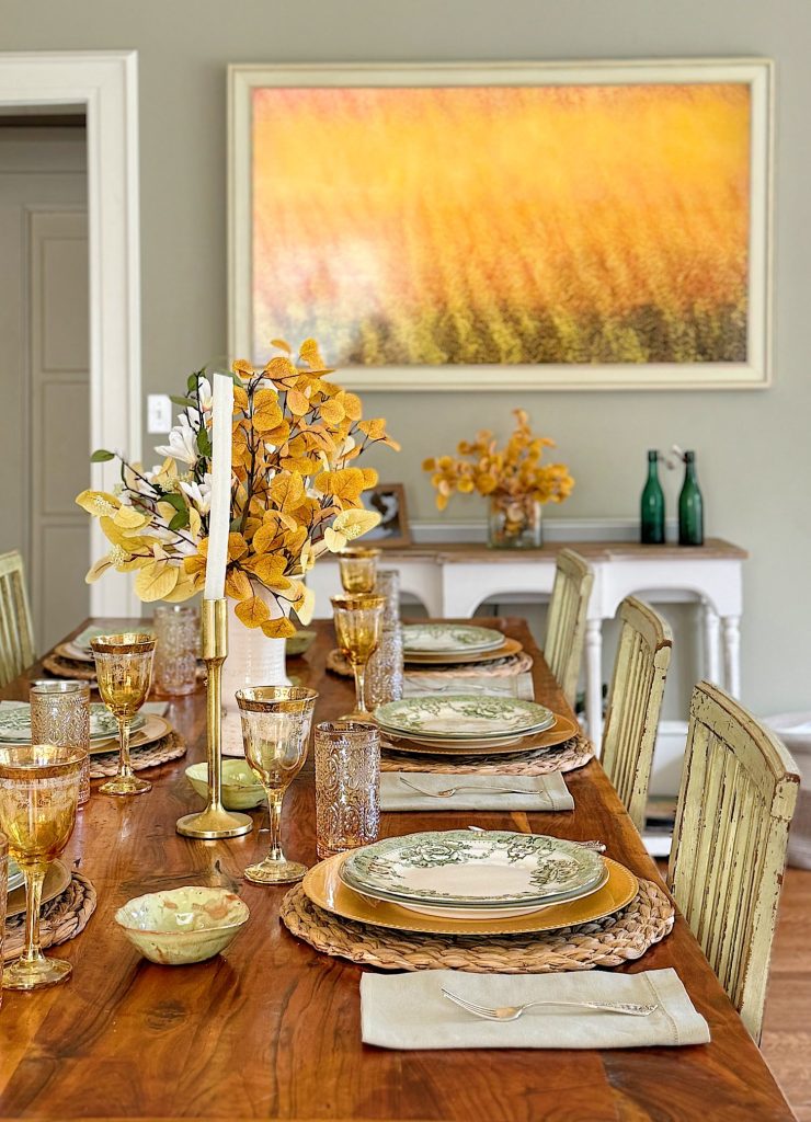 A dining table set with plates, glasses, and a centerpiece of yellow foliage. In the background, a matching wall art and a side table with bottles and more yellow decorations are visible.