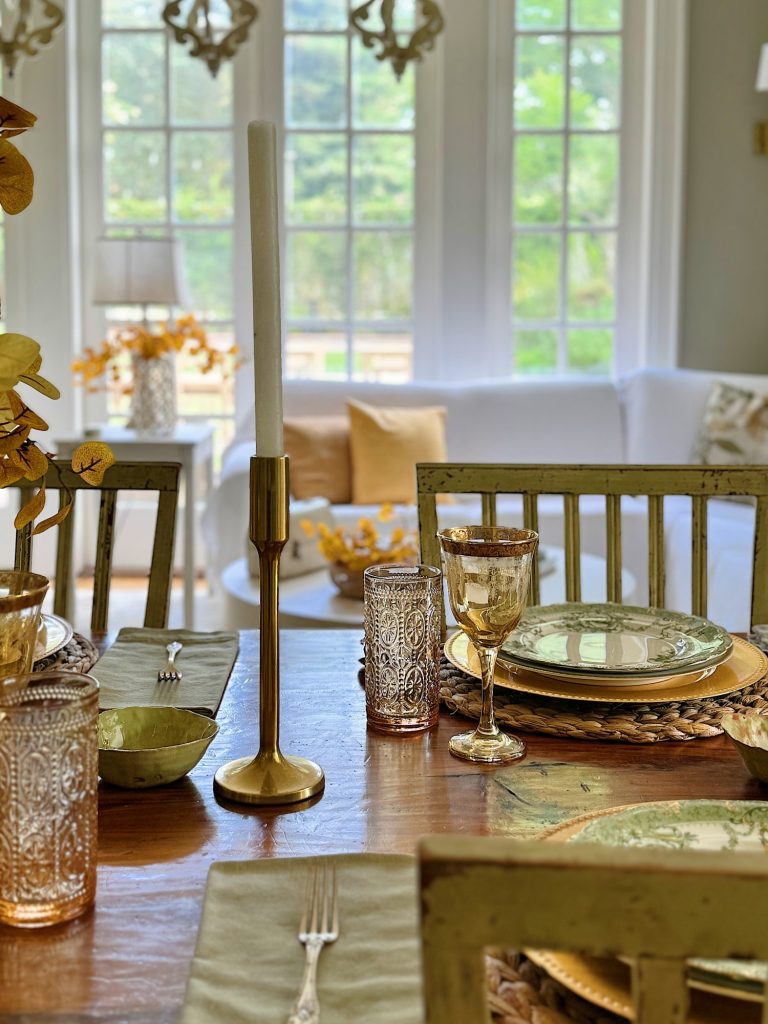 A dining table set with plates, glasses, silverware, and a candlestick in a sunlit room with a white couch and large windows in the background.