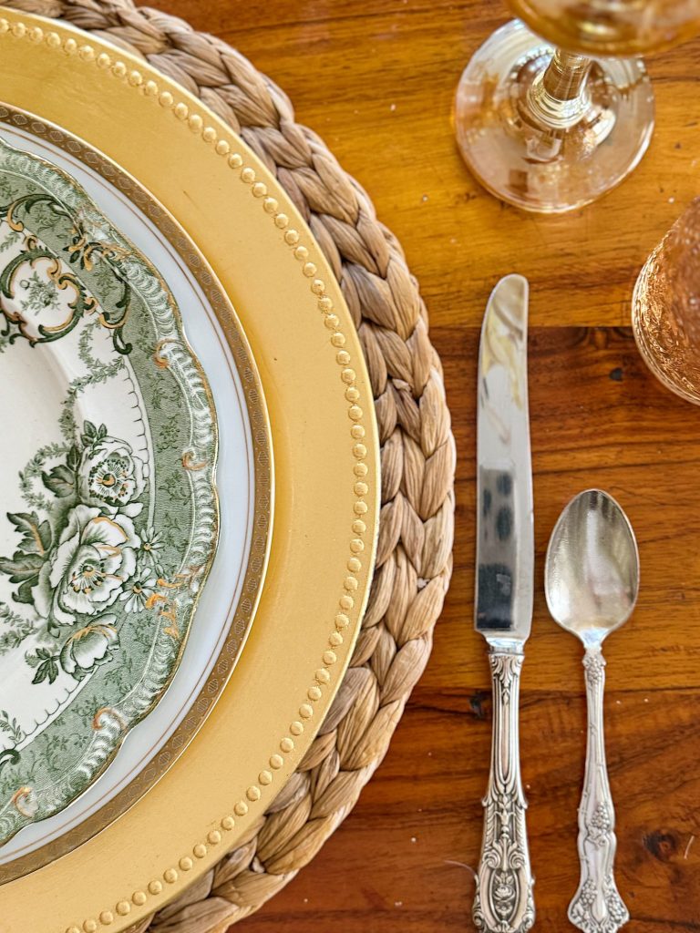 Close-up of a table setting with a decorative plate, golden charger, woven placemat, knife, and spoon, alongside a clear and a pink glass on a wooden table.