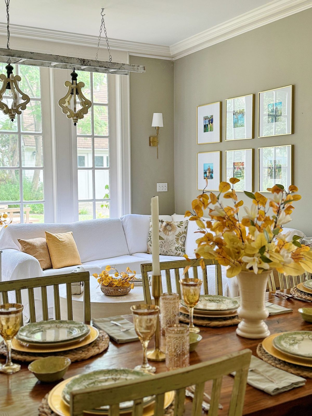 A well-lit living and dining room with a white sofa, yellow pillows, and a wooden dining table set for a meal. The table has golden accents, and there are framed photos and a vase with flowers on the wall.