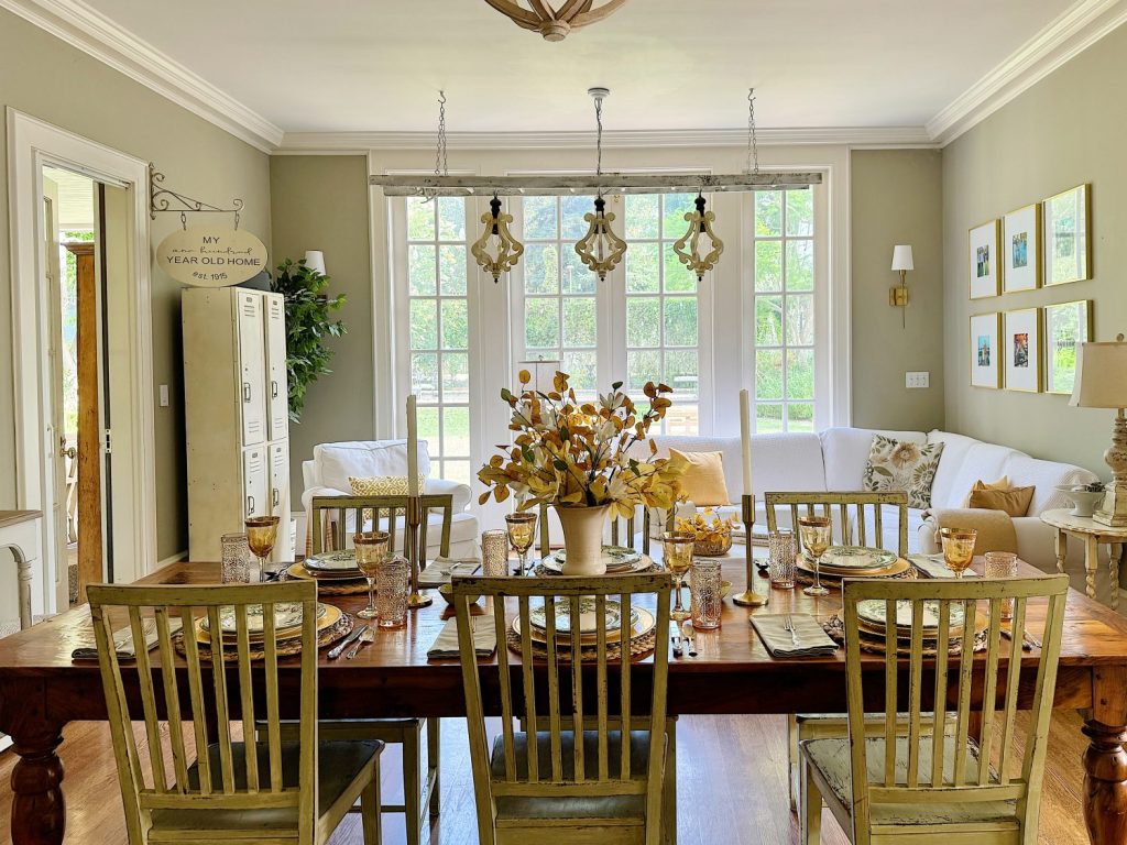 A bright, elegant dining room with a wooden table set for eight. Large windows in the background allow natural light to fill the space, which is decorated with plants and framed pictures.