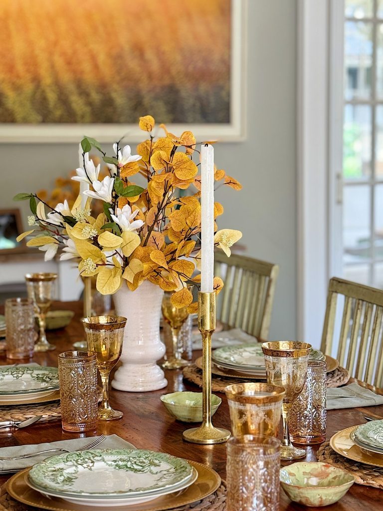 A dining table set with ornate plates, golden glasses, candlesticks, and a centerpiece of yellow and white flowers in a vase. A large framed picture of a field hangs on the wall behind it.