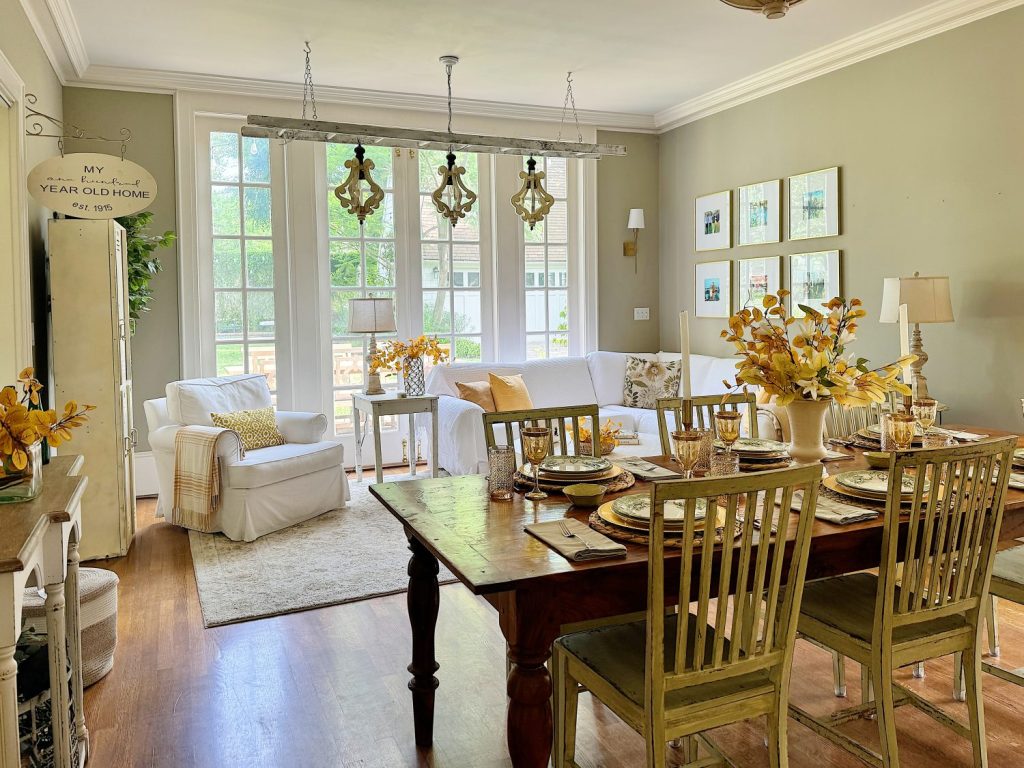 Image of a bright, cozy dining and living area with wooden floors, a large wooden table set for a meal, green chairs, a white sofa, an armchair, and large windows allowing natural light to fill the room.