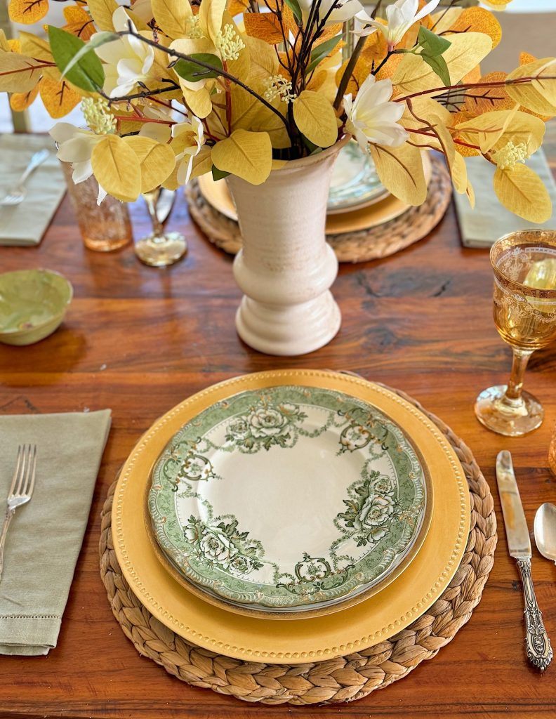 A dining table setting includes a green and white floral plate on a yellow charger, a woven placemat, a napkin, silverware, a goblet, and a floral centerpiece in a white vase.