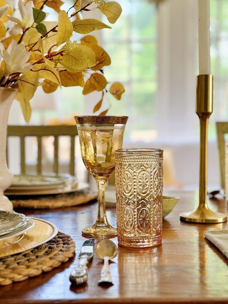 A dining table setting with ornate glassware, cutlery, a wreath placemat, and a vase of yellow leaves. A gold goblet and glass are placed beside a candlestick on a wooden table.