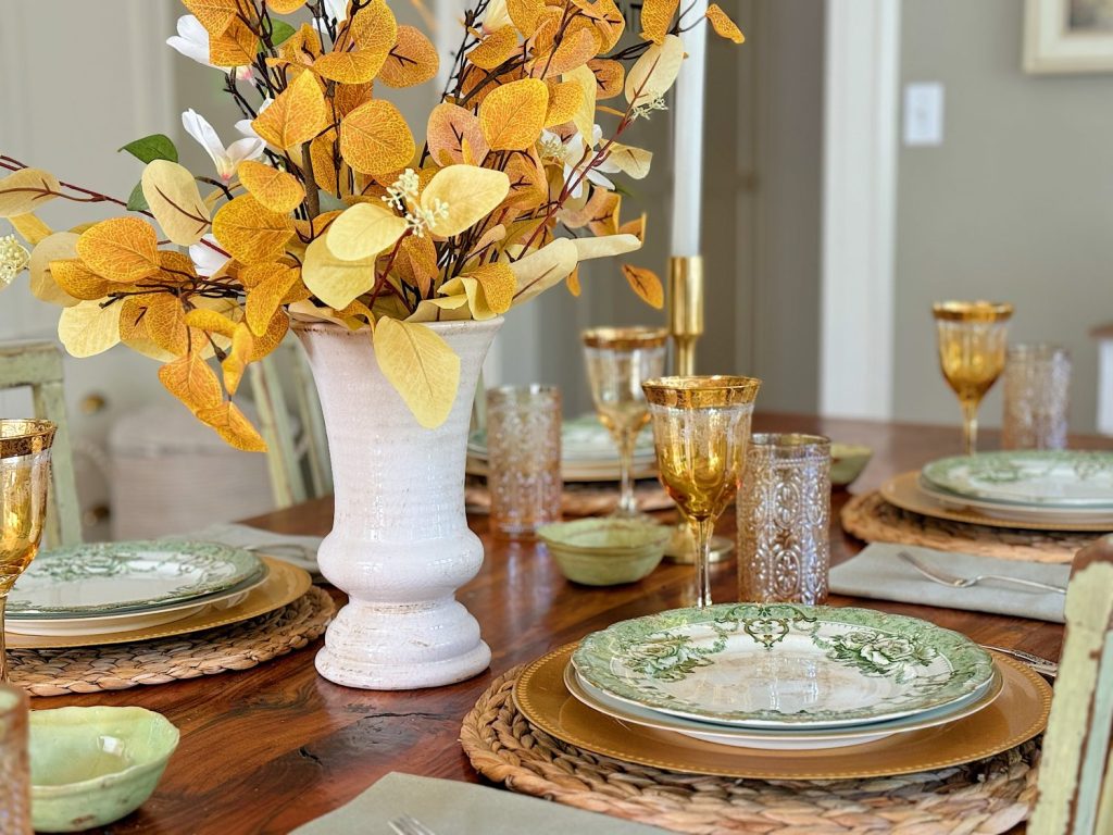 A dining table is set with green and beige plates, amber-colored glasses, and a centerpiece of yellow and white leaves in a white vase. Woven placemats and beige napkins complement the setting.