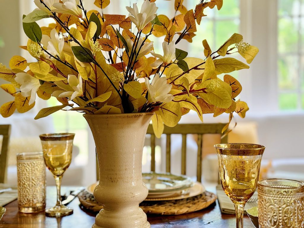 A beige vase with white and yellow flowers sits on a wooden table set with glassware and plates in a bright room.