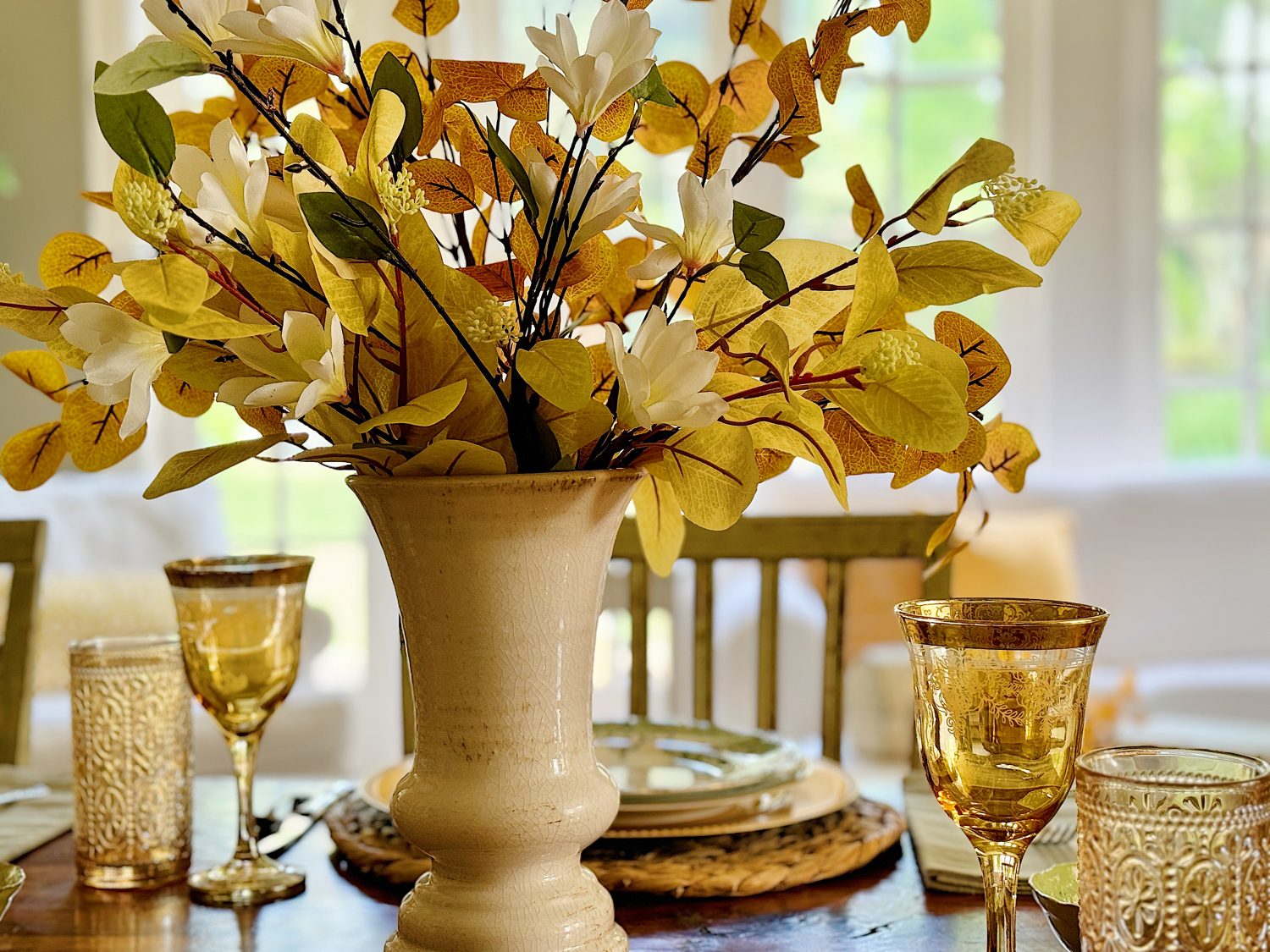 A beige vase with white and yellow flowers sits on a wooden table set with glassware and plates in a bright room.