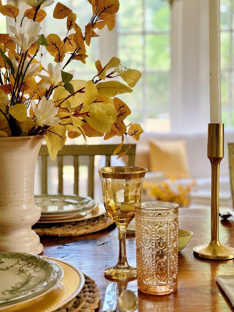 A dining table adorned with a floral centerpiece, gold-rimmed plates, ornate drinking glasses, and a gold candle holder. The table is set in a bright room with large windows in the background.