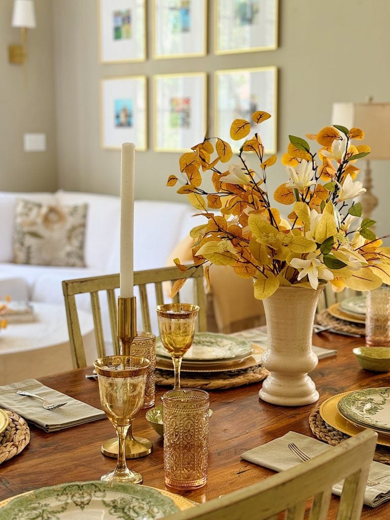 A dining table is set with green-patterned plates, gold-rimmed glasses and cutlery, and a centerpiece of yellow flowers. A white sofa and framed pictures are in the background.