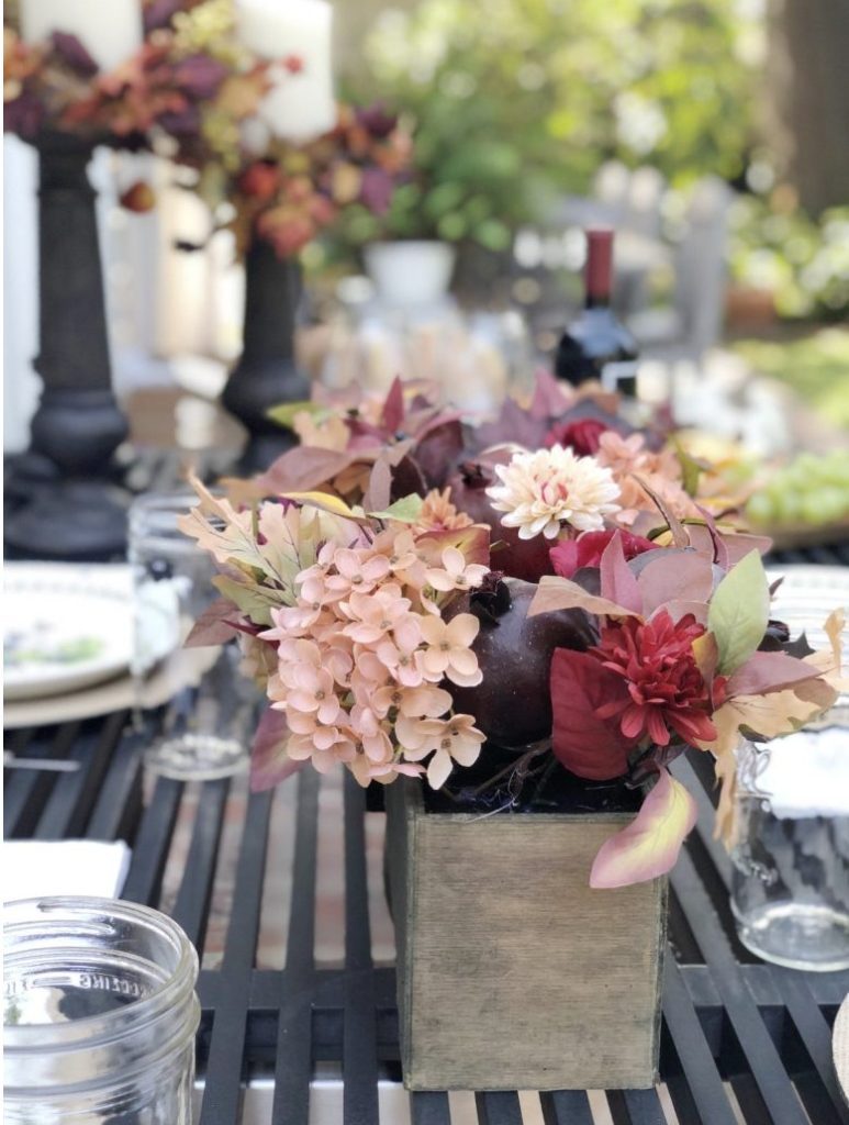 A wooden box filled with red and pink flowers and foliage sits on an outdoor table set for dining, with glass jars and a bottle of wine in the background.