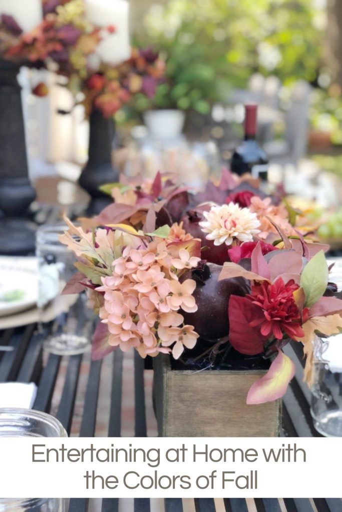 A fall-themed centerpiece with flowers and leaves in a wooden container on a dining table set outdoors. Wine bottles and glasses are seen in the background. Text reads: "Entertaining at Home with the Colors of Fall.