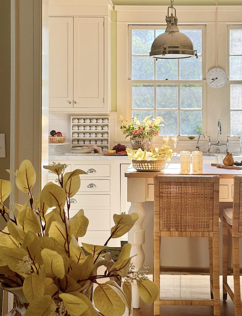 A bright kitchen with white cabinets, a large window, a wooden island and wicker stools. A metallic pendant light hangs above the island, and yellow flowers are in the foreground.