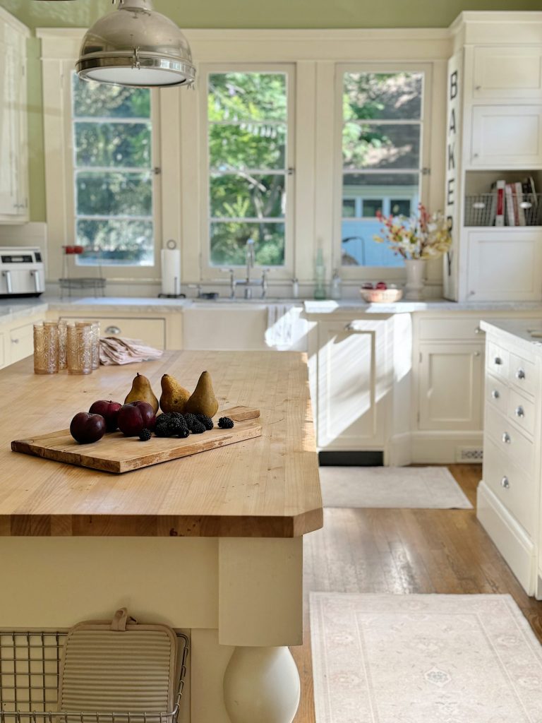 Bright kitchen with large windows, a wooden island countertop, and a cutting board with pears and plums in the foreground. Cabinets, sink, and appliances are in the background.