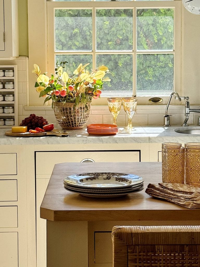 A kitchen counter displays plates, glasses, and folded napkins. A vase with flowers and dishes of fruit are near a sink with a window above it showcasing greenery outside.