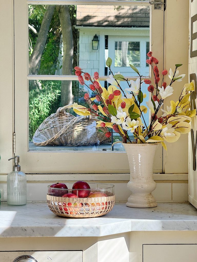 A kitchen windowsill with a vase of flowers, a bowl containing red apples, a soap dispenser, and a view of a wicker basket and greenery outside.