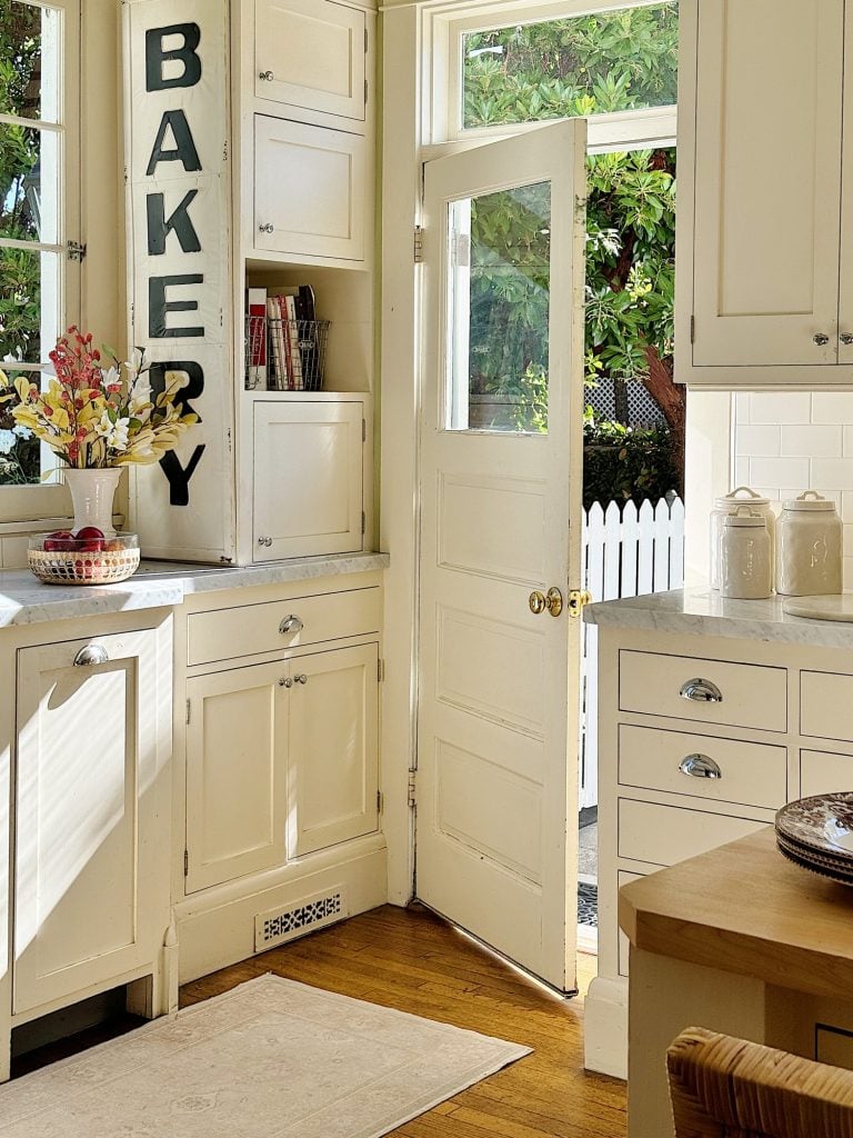 A bright kitchen with white cabinets, a sign reading "BAKERY," and an open door leading outside. The countertop has a bowl of fruit and jars, with a cozy and inviting ambiance.