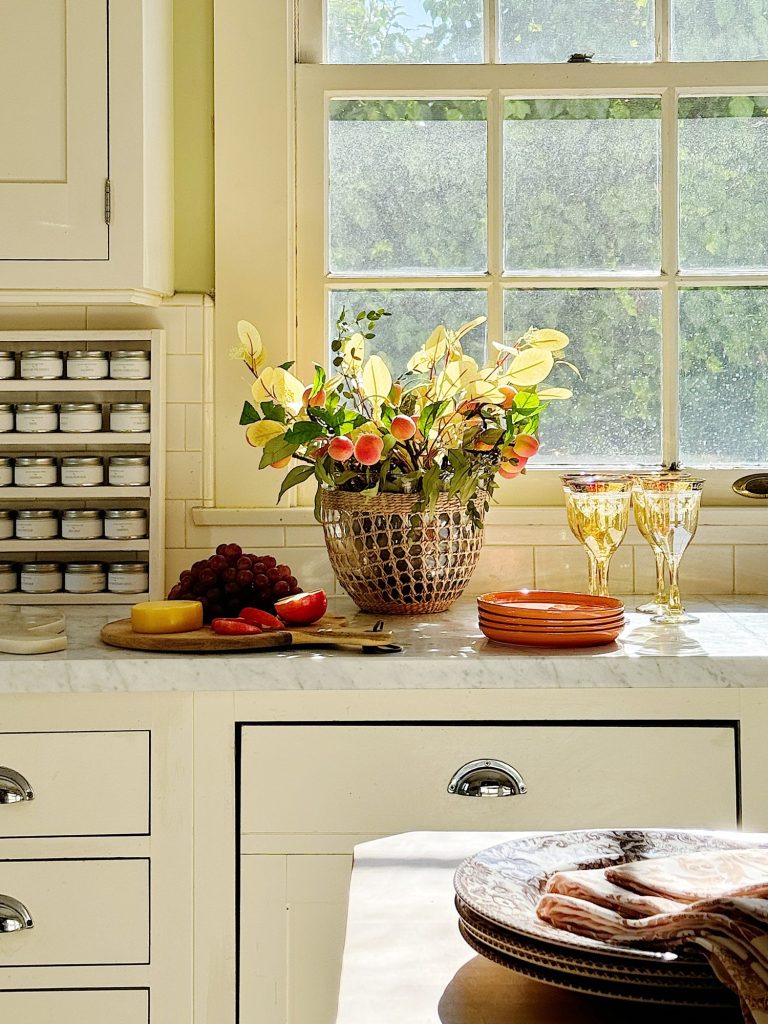 A sunlit kitchen counter features a basket of artificial fruit and flowers, a wooden platter with fruit and cheese, stacked orange plates, and glassware in front of a window with a greenery view outside.