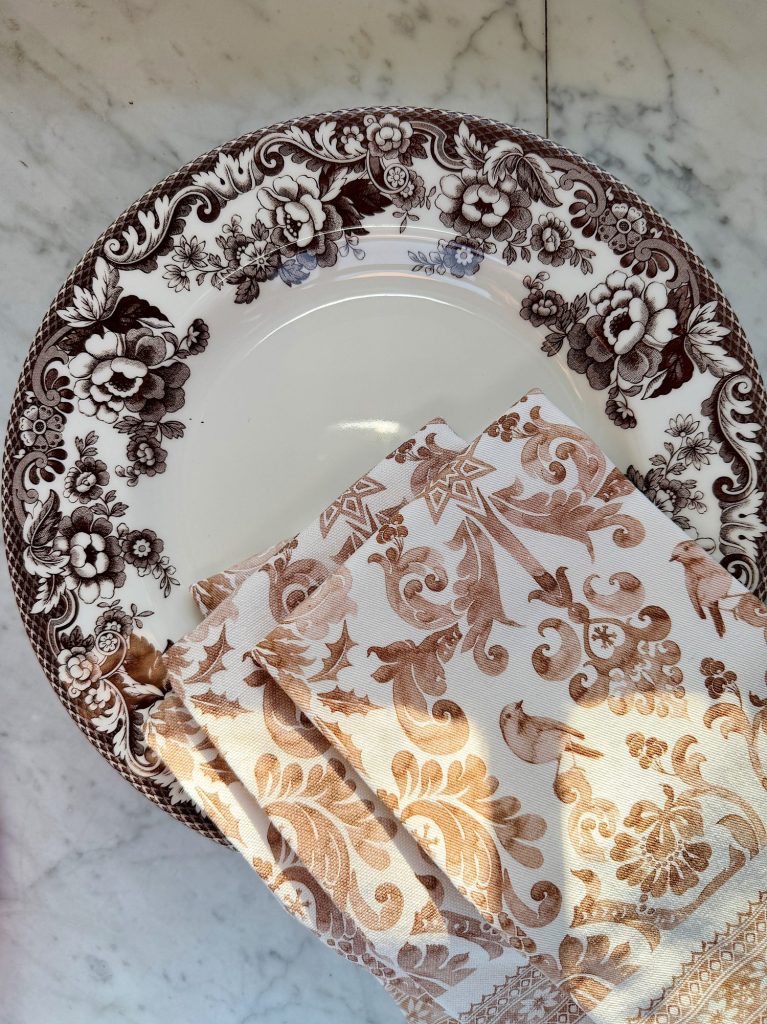 A floral-patterned plate and a folded, beige, patterned cloth napkin are placed on a marble surface.